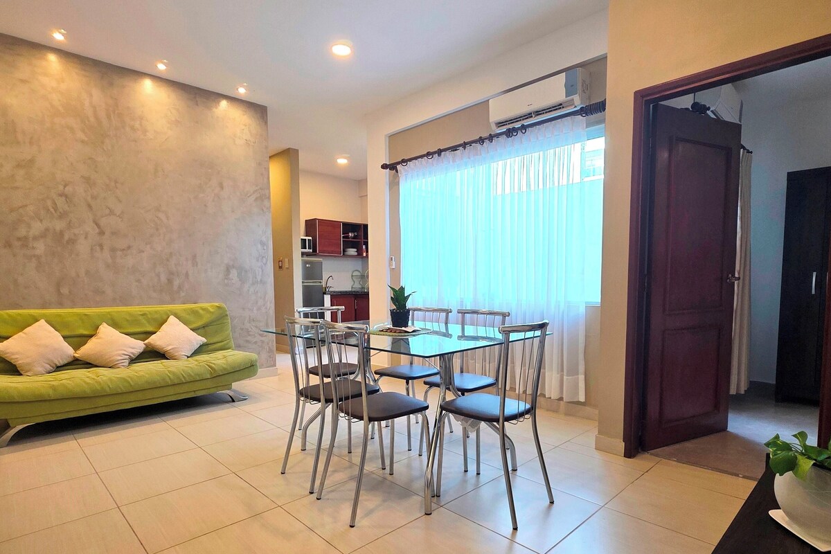 A living room with a green sofa, a glass dining table with chairs, a kitchen area, and a large window with curtains.