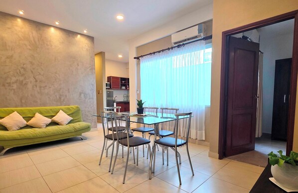 A living room with a green sofa, a glass dining table with chairs, a kitchen area, and a large window with curtains.