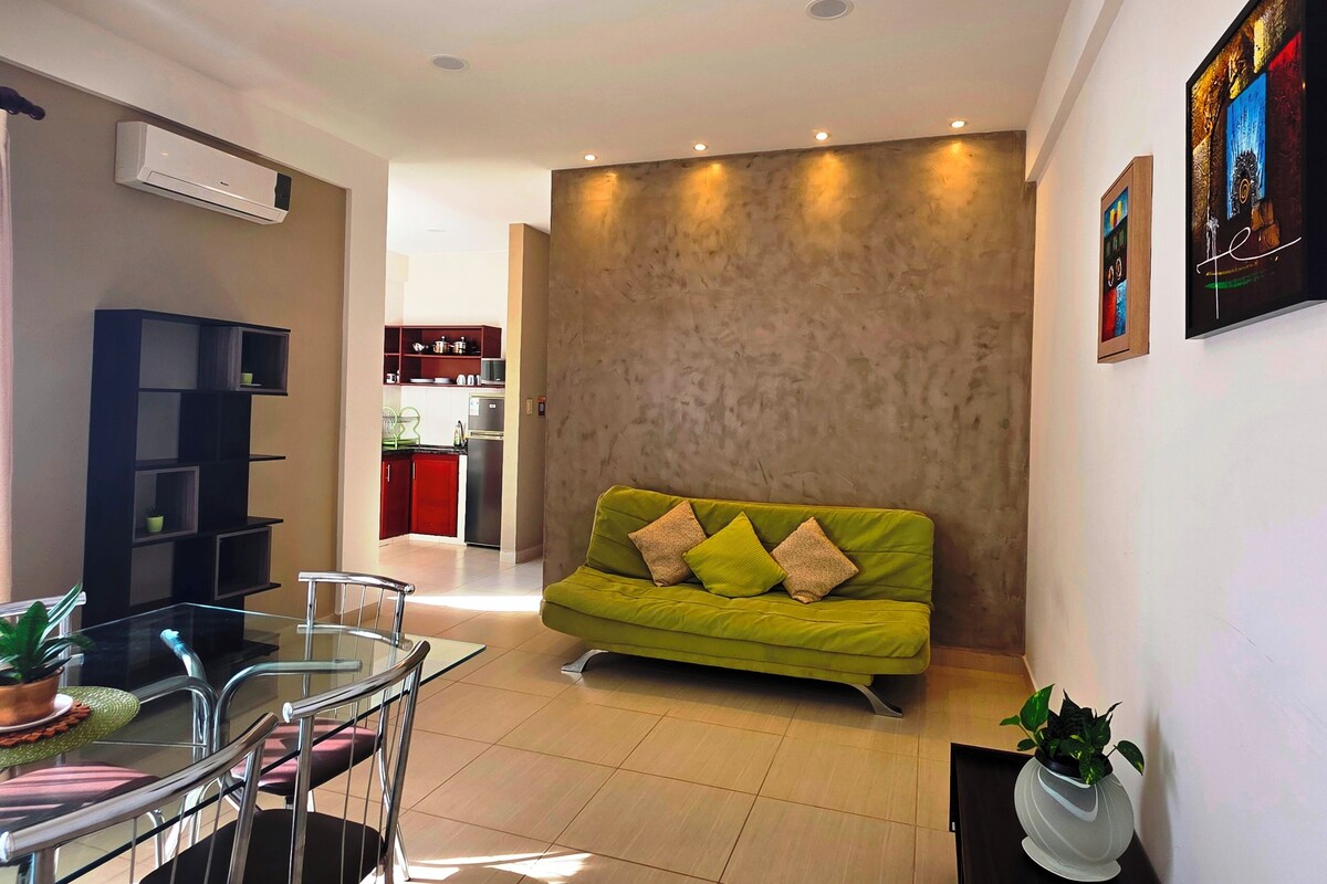 A living room with a green sofa, a glass dining table with chairs, a kitchen in the background, and a wall-mounted air conditioner.