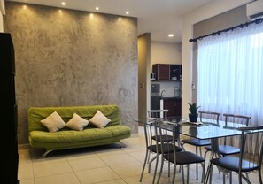 Sala de estar con sofá verde, mesa de comedor de vidrio con sillas, área de cocina al fondo y un ventanal con cortinas.