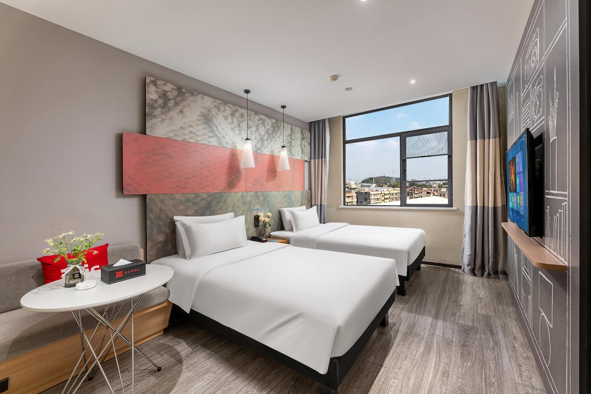 Habitación de hotel con dos camas, una mesita con un florero, un televisor y un ventanal con vistas a la ciudad.