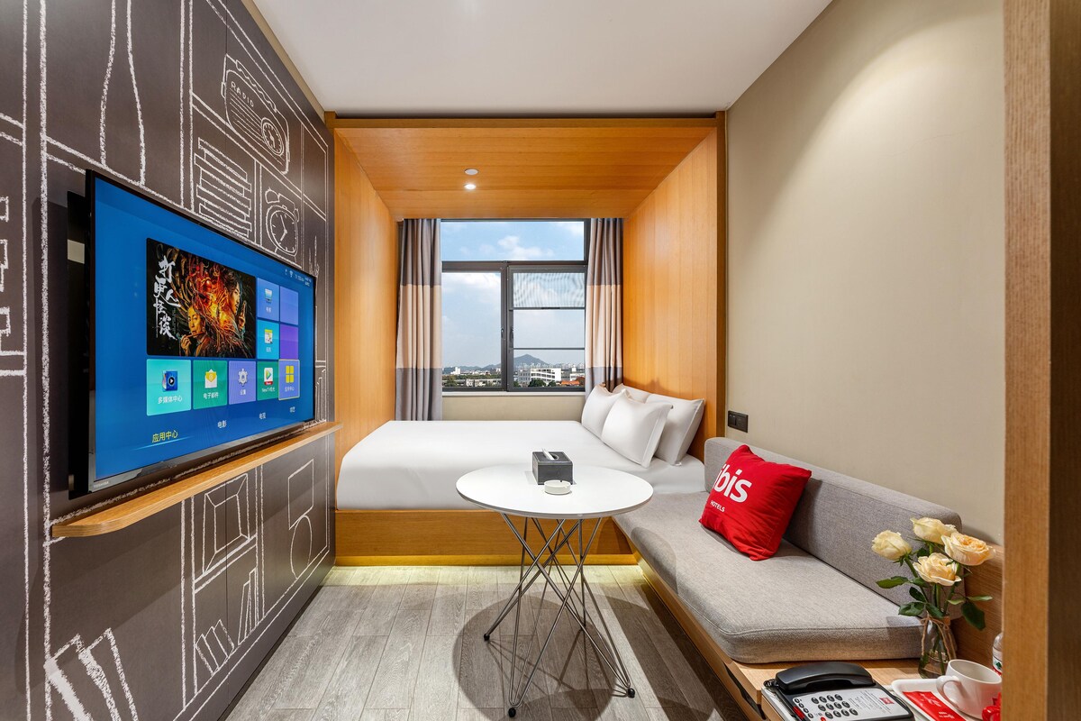 A hotel room with a bed, a sofa, a TV mounted on the wall, a small table, and a window with a city view.