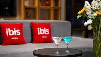 A room with a grey sofa, two red pillows with 'ibis' branding, a small round table with two martini glasses, and a vase with flowers.