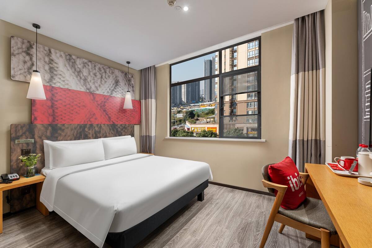 Habitación de hotel con cama, escritorio, ventana y vista a la ciudad, además de una almohada roja.