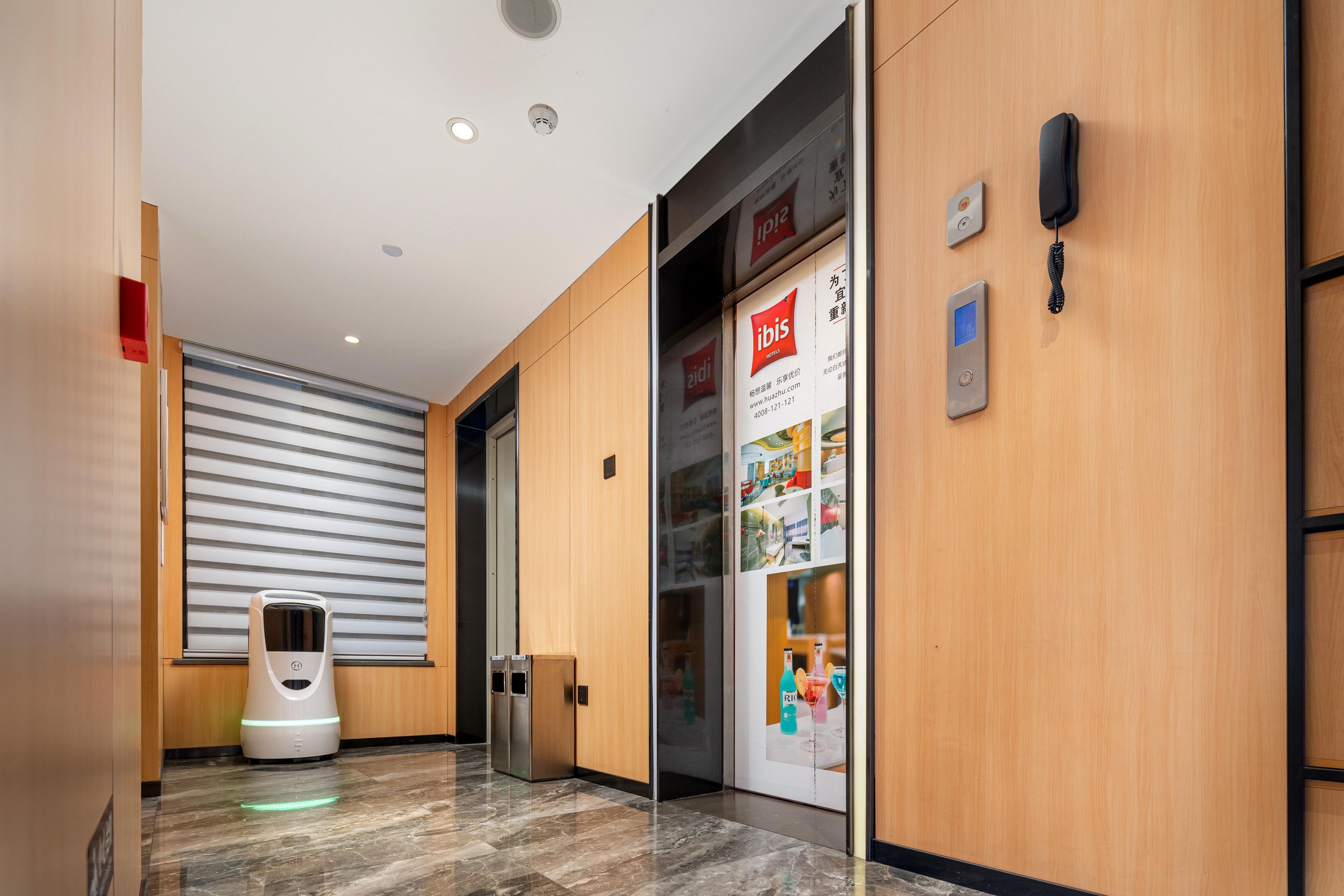 A hotel corridor with a self-service kiosk, a glass door with a poster, and a phone on the wall.