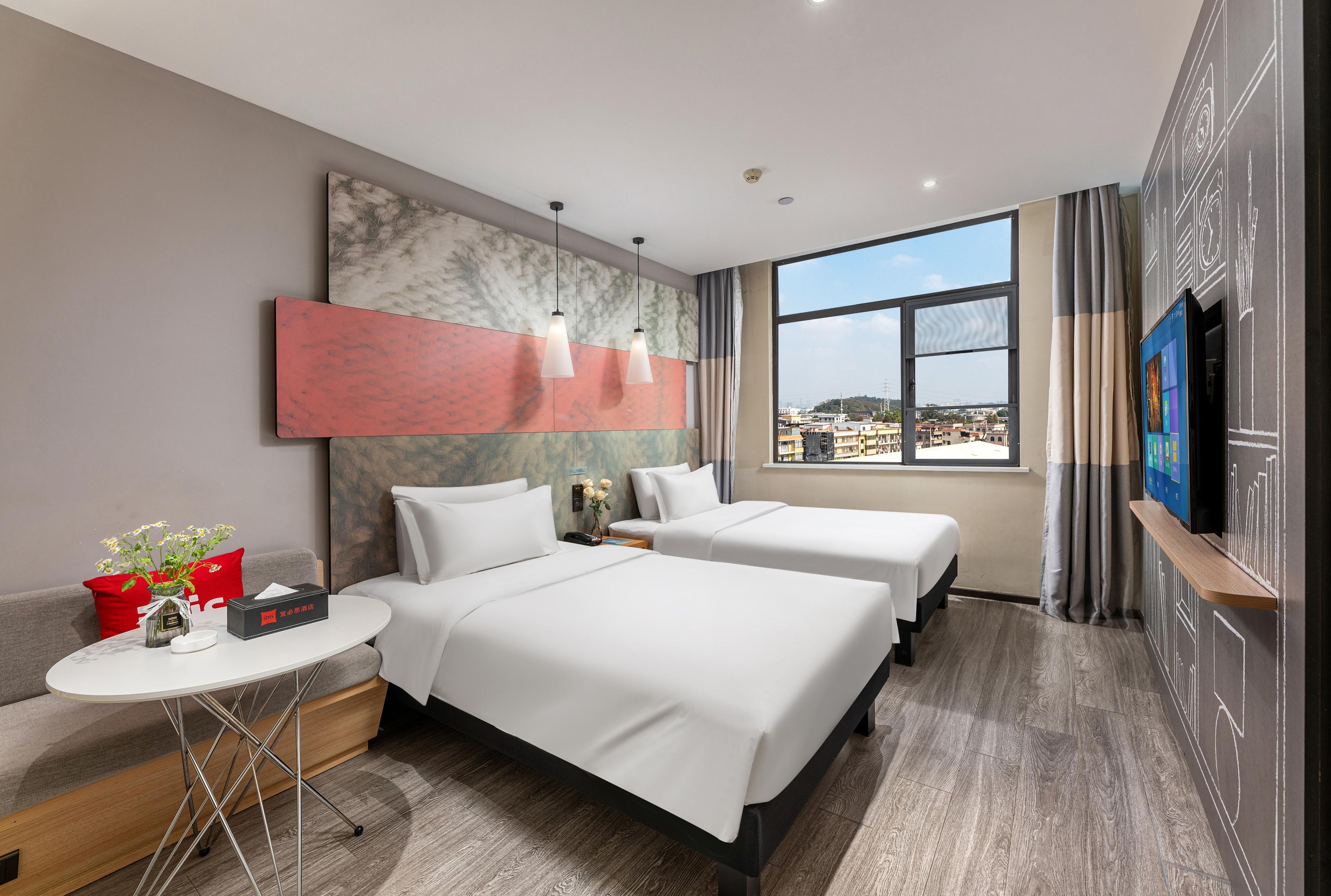 Hotelkamer met twee bedden, een tafeltje met een vaas, een televisie en een groot raam met uitzicht op de stad.