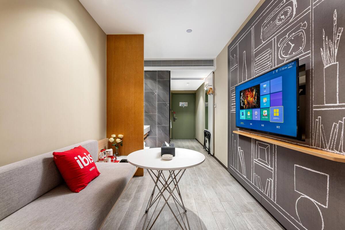 Una habitación de hotel moderna con televisor de pantalla plana, una mesa redonda, un sofá con un cojín rojo y una pared con dibujos al estilo de pizarra.