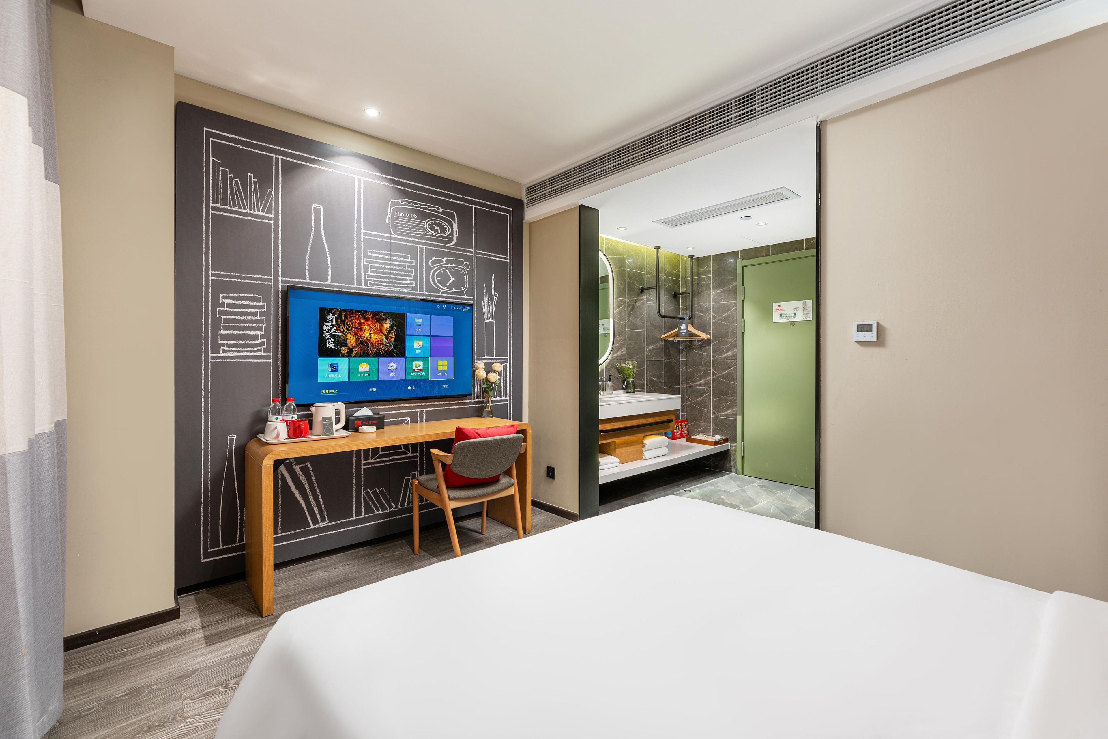 A hotel room with a bed, a desk with a computer, a chair, a television, and a bathroom with a mirror and towel rack.