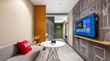 A modern hotel room with a flat-screen TV, a round table, a sofa with a red pillow, and a wall with chalkboard-style drawings.
