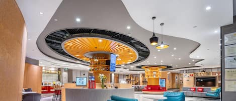 Un hall d’hôtel moderne doté d’un plafond incurvé, d’un bar central, de différents espaces aménagés avec des sièges et d’un comptoir de réception.