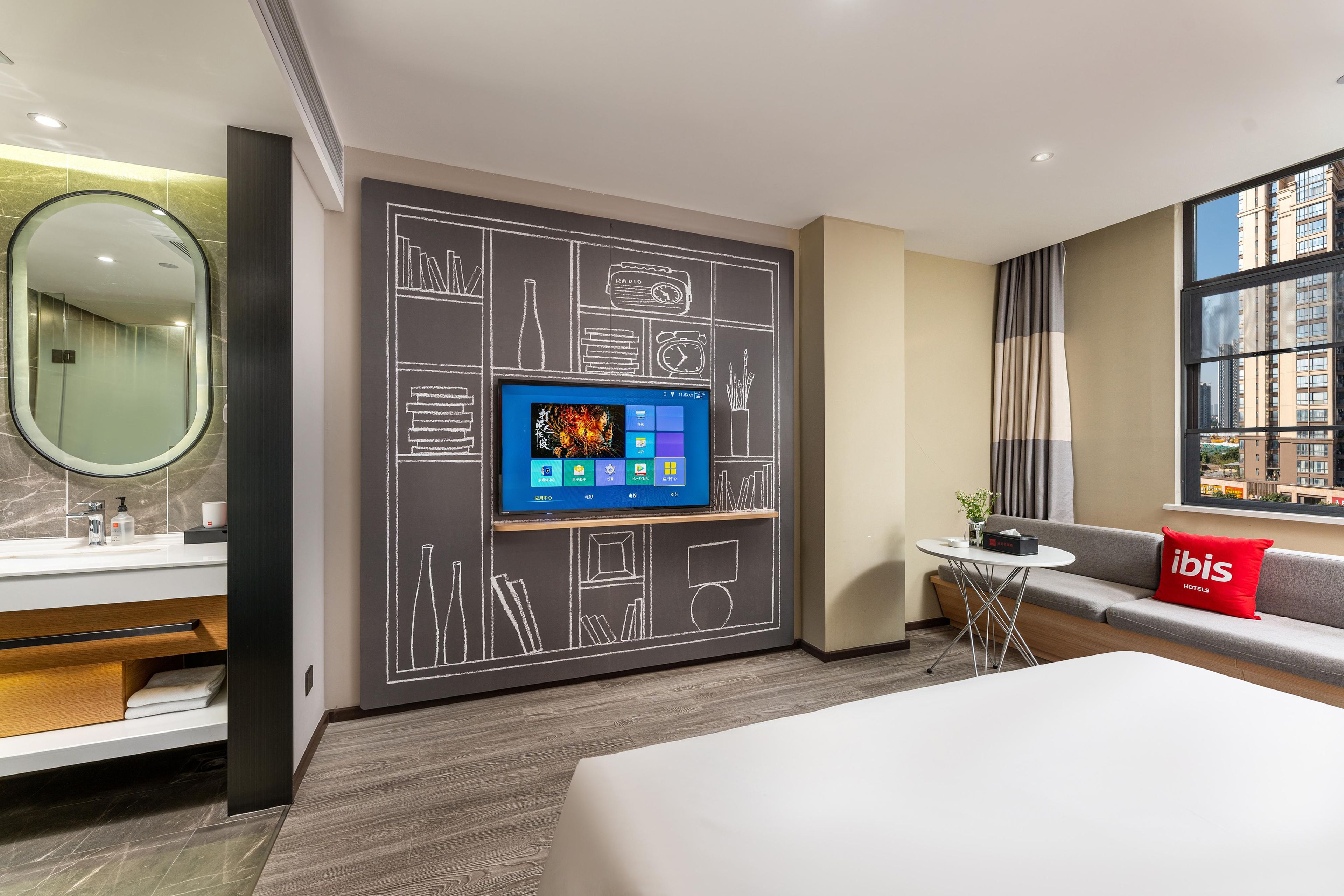 A modern hotel room with a large bed, a TV mounted on the wall, a small round table, and a sofa with a red pillow.