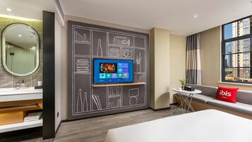 A modern hotel room with a large bed, a TV mounted on the wall, a small round table, and a sofa with a red pillow.