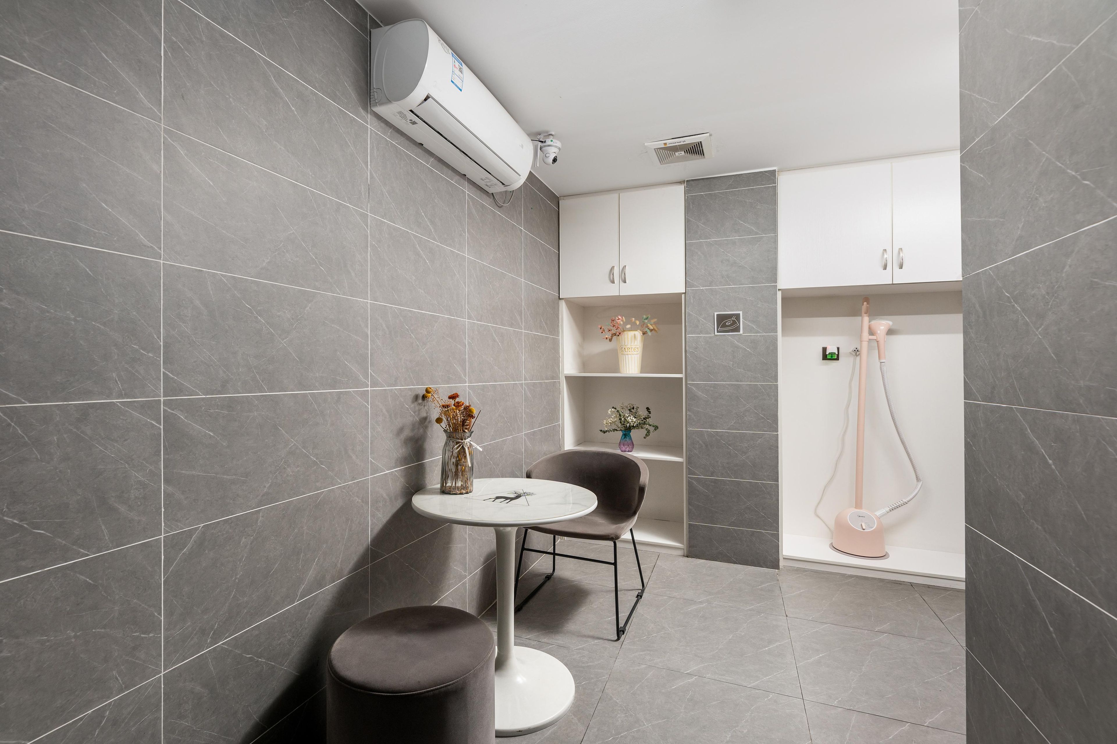 A modern bathroom with grey tiles, a white air conditioning unit, a small round table with a vase, a chair, and a shower area.