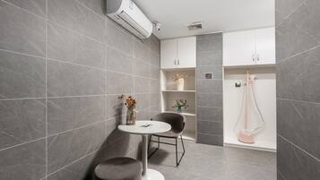 A modern bathroom with grey tiles, a white air conditioning unit, a small round table with a vase, a chair, and a shower area.