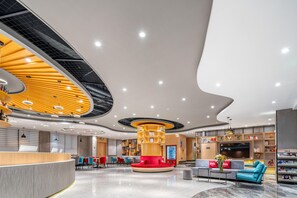 A modern lobby with a curved reception desk, a central seating area with red and blue chairs, and a large bookshelf. - Ibis Hotel Foshan Shunde Jing Long Subway Station (Foshan)