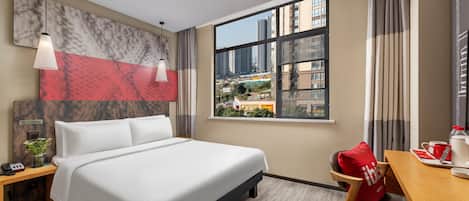 A hotel room with a bed, a desk, a window with a city view, and a red pillow.