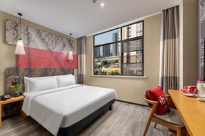 Habitación de hotel con cama, escritorio, ventana y vista a la ciudad, además de una almohada roja.