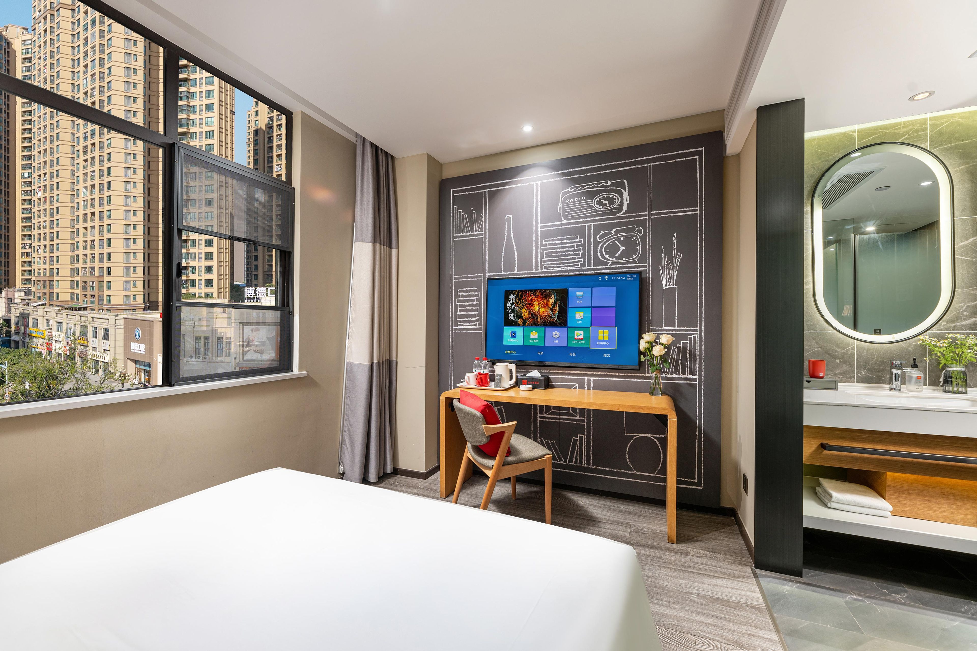A hotel room with a large window, a desk with a computer, a chair, a bed, and a bathroom with a mirror and sink.