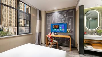 A hotel room with a large window, a desk with a computer, a chair, a bed, and a bathroom with a mirror and sink.