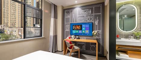 A hotel room with a large window, a desk with a computer, a chair, a bed, and a bathroom with a mirror and sink.