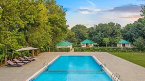 Una piscina rectangular con superficie de agua azul cristalina, rodeada por un solárium empedrado. Hay sillones de playa con sombrillas en el lado izquierdo, y una hilera de edificios con techos verdes al fondo.