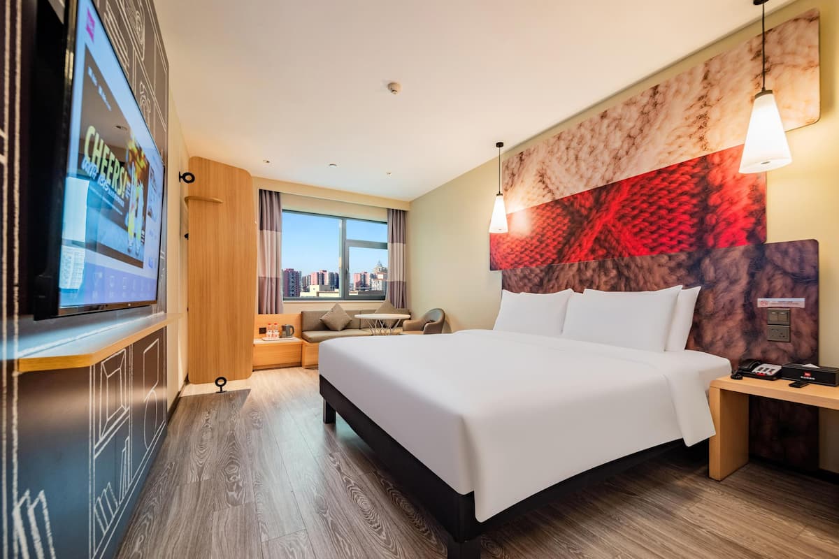 Habitación de hotel con una cama grande, televisor en la pared, vista a la ciudad y cabecera moderna.