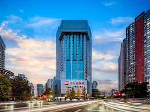 A modern high-rise building with a prominent glass facade, surrounded by other tall buildings, and a busy urban street with light trails indicating traffic. - Ibis Chengdu Kuanzhai Alley Center Hotel (Chengdu)