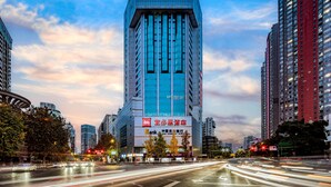 A modern high-rise building with a prominent glass facade, surrounded by other tall buildings, and a busy urban street with light trails indicating traffic. - Ibis Chengdu Kuanzhai Alley Center Hotel (Chengdu)