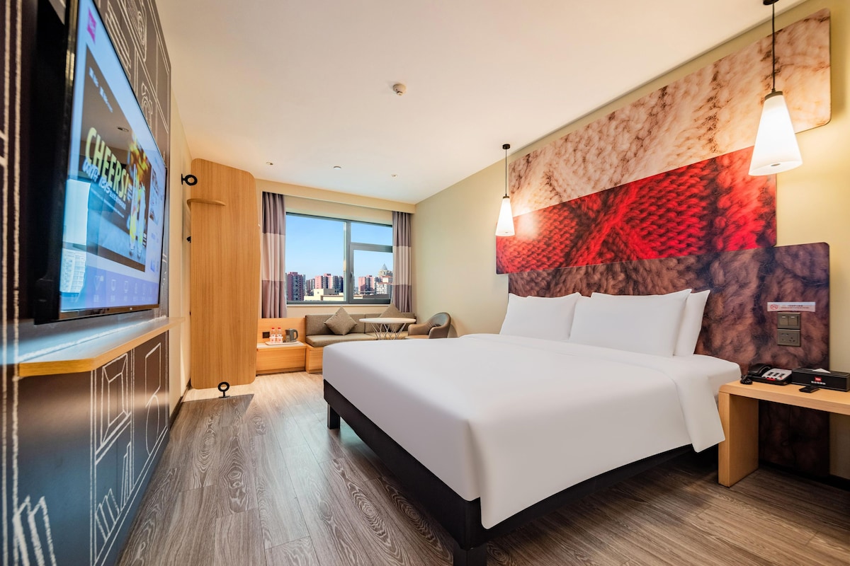 A hotel room with a large bed, a TV on the wall, a city view, and a modern headboard.