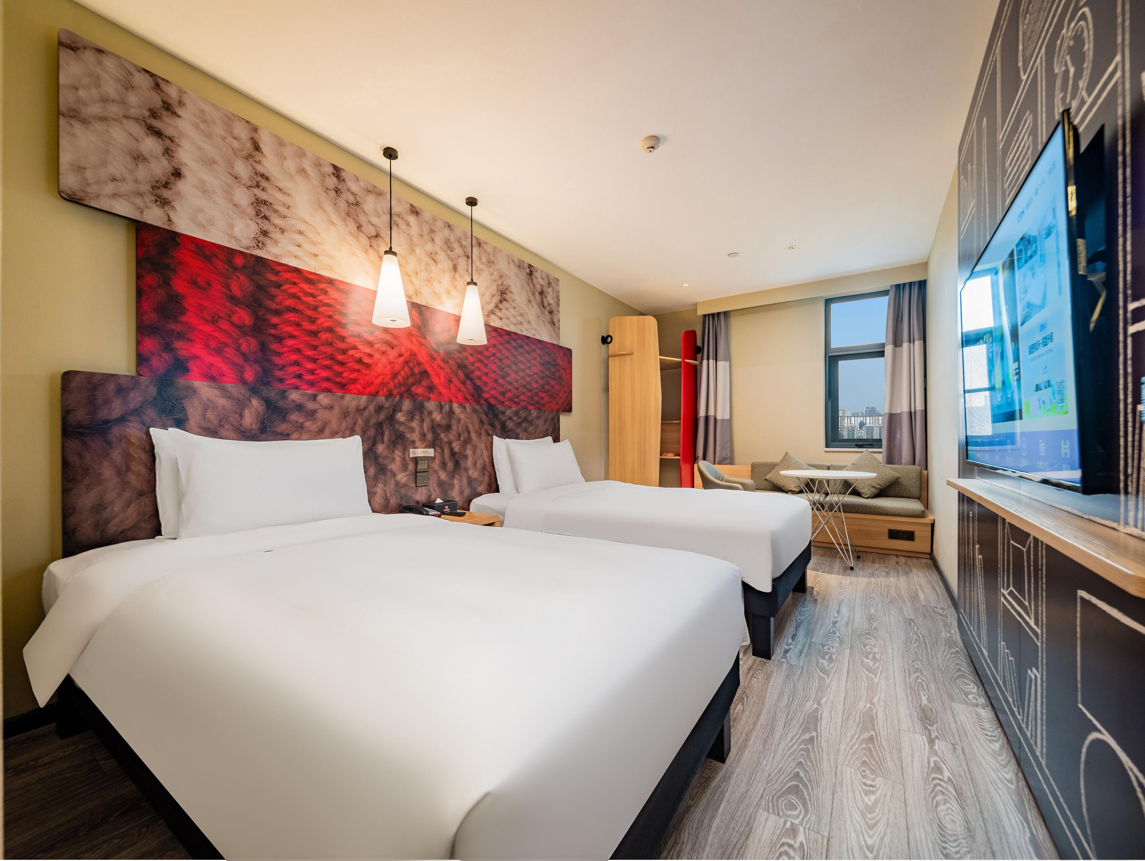 A hotel room with two beds, a TV, a desk, and a view of the city.