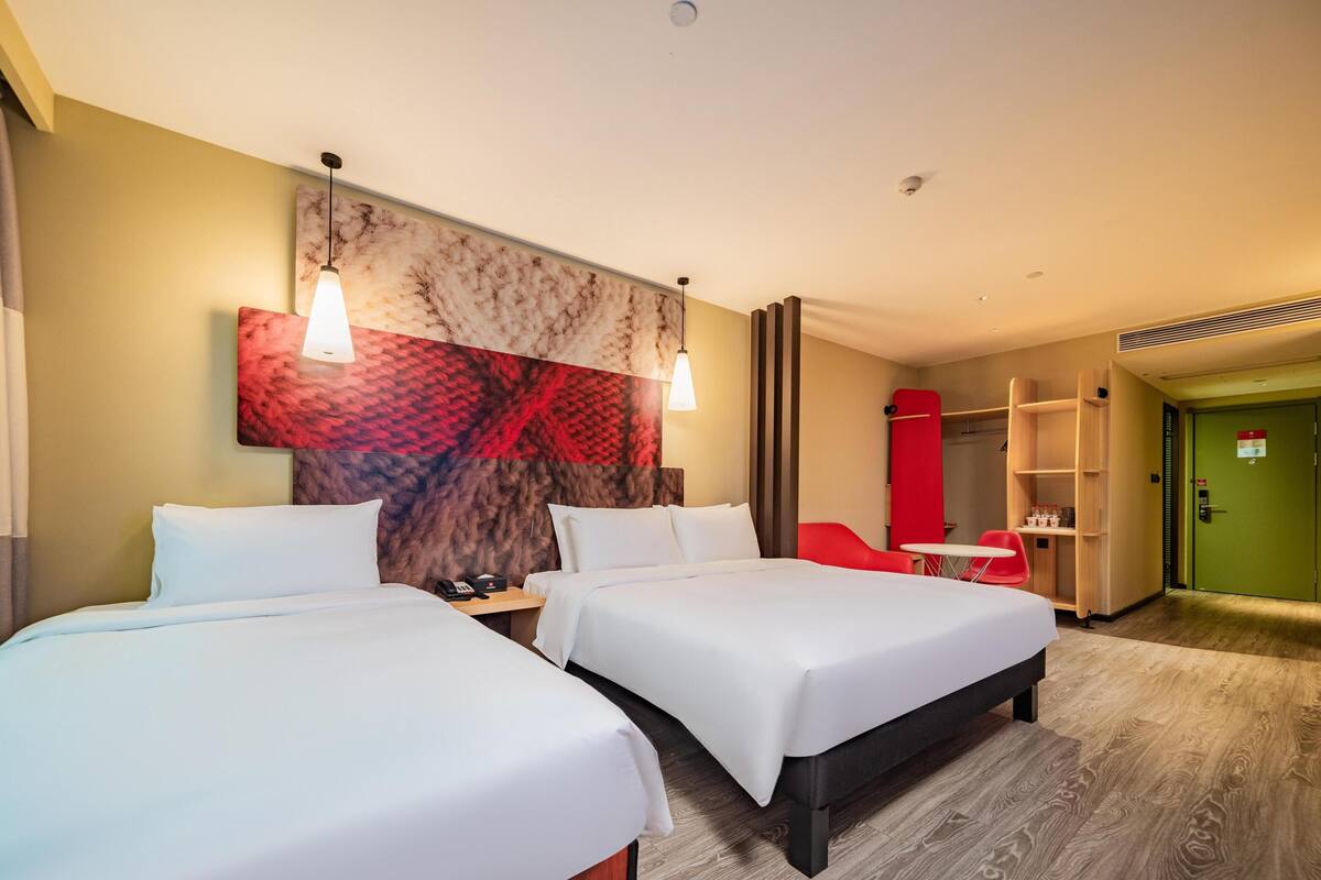 Habitación de hotel con dos camas, una silla roja, una mesita pequeña y una puerta verde.