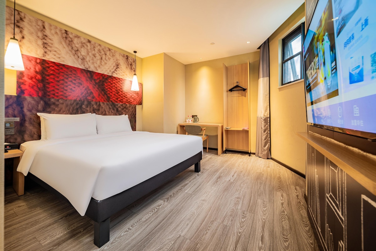 A hotel room with a bed, bedside table, lamp, TV, and a wall with a red and beige pattern.