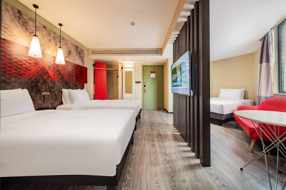 Una habitación de hotel con dos camas, un televisor, una silla roja y una pequeña mesa.