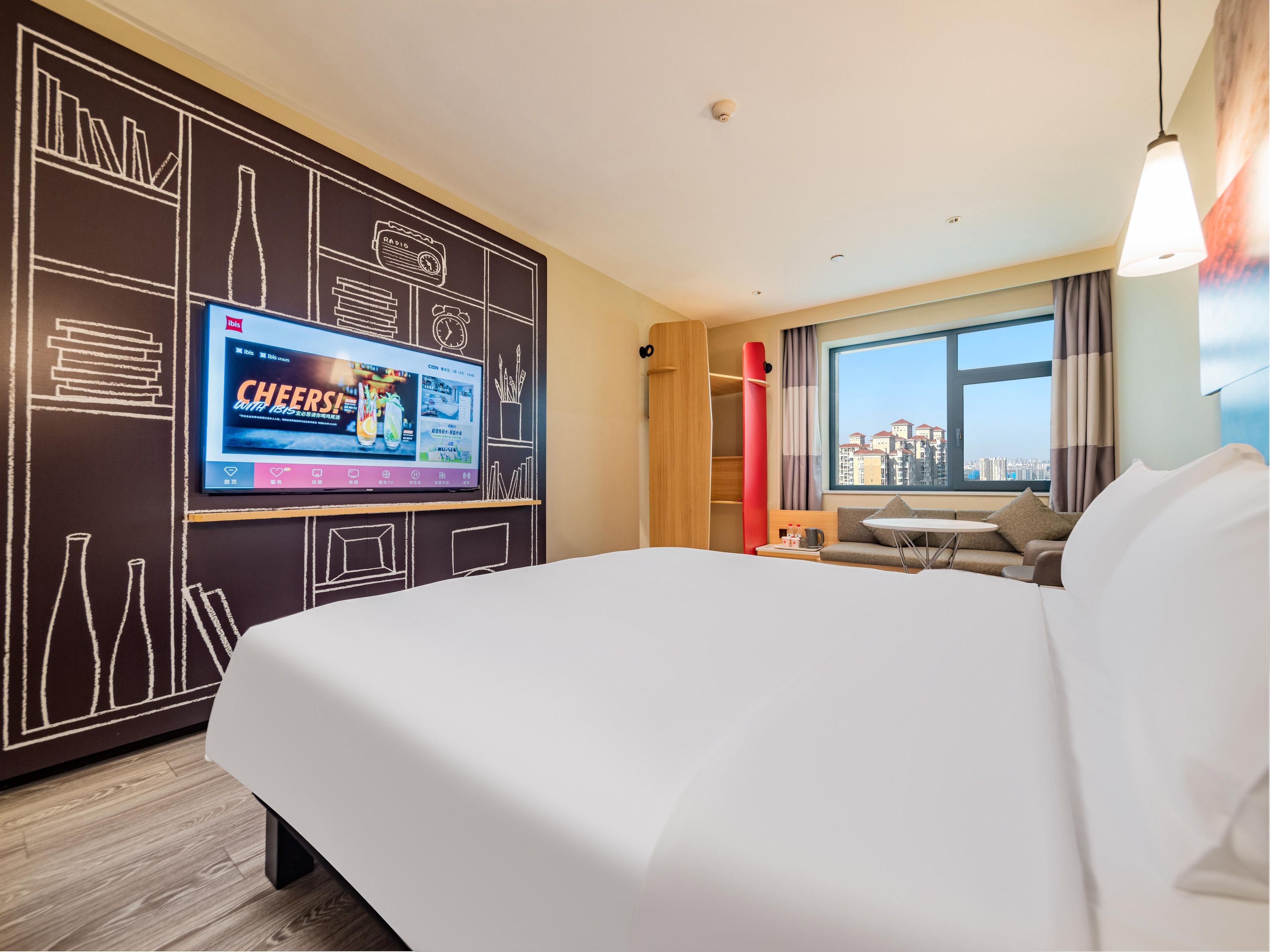 A hotel room with a large bed, a television displaying a commercial, a city view through the window, and a modern design with chalkboard walls.