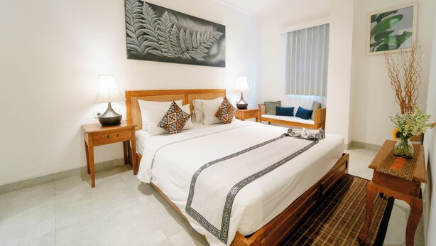 A hotel room with a large bed, two bedside tables with lamps, a wooden headboard, a large fern artwork on the wall, a window with curtains, and a small table with a vase of flowers. - Umalas Suites Bali (Kerobokan)