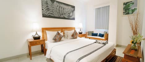 Habitación de hotel con una cama grande, dos mesitas de noche con lámparas, un cabecero de madera, una obra de arte con helechos en la pared, una ventana con cortinas y una pequeña mesa con un jarrón de flores.
