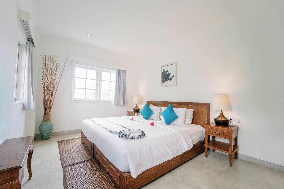 A bedroom with a large bed, bedside lamps, a wooden nightstand, a desk, a vase with branches, and a window with curtains.