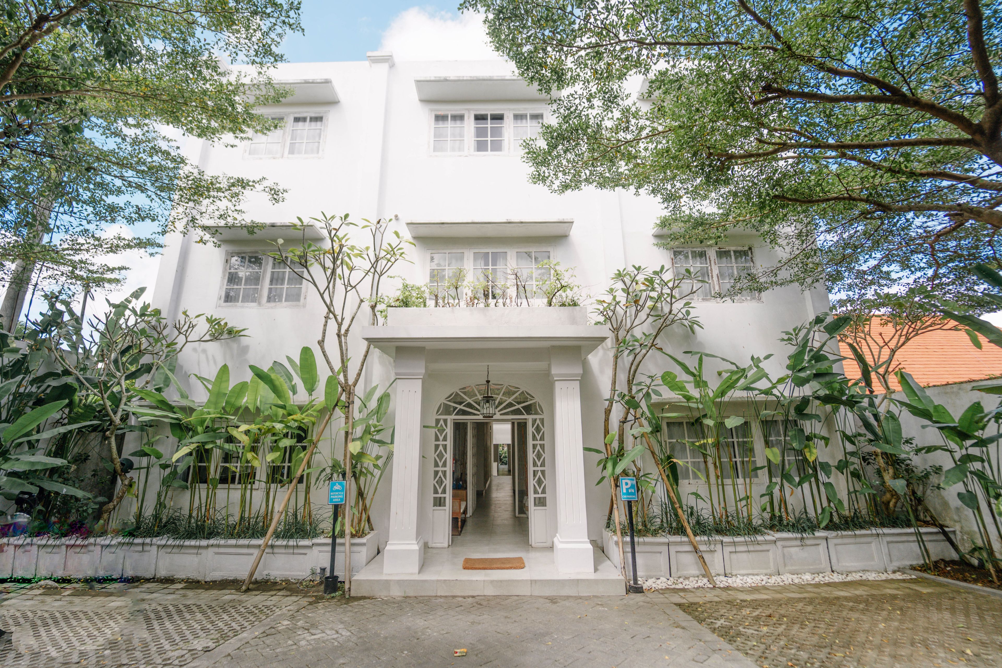 A white building with a central entrance, surrounded by greenery and a paved area.
