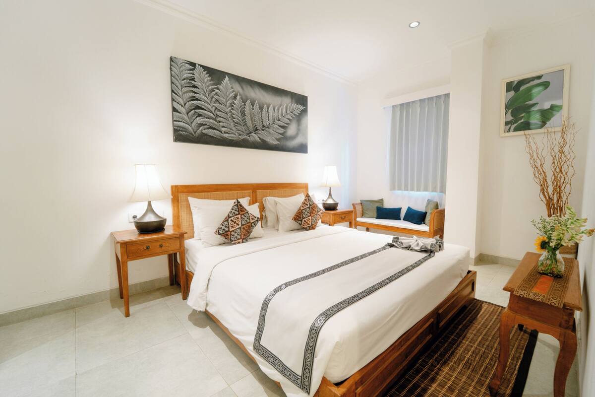 A hotel room with a large bed, two bedside tables with lamps, a wooden headboard, a large fern artwork on the wall, a window with curtains, and a small table with a vase of flowers.