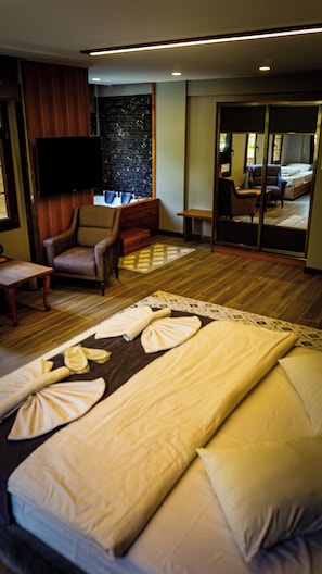 Habitación de hotel con cama, televisor, zona de estar con una silla y una mesa, y un espejo grande.