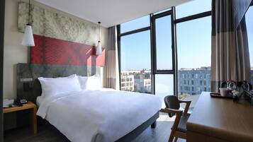 A hotel room with a large bed, a desk, a chair, a nightstand, and a view of the city through large windows.