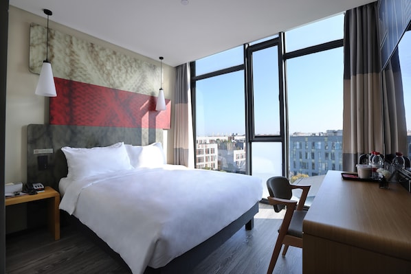 Habitación de hotel con una cama grande, un escritorio, una silla, una mesita de noche y vistas a la ciudad a través de amplios ventanales.