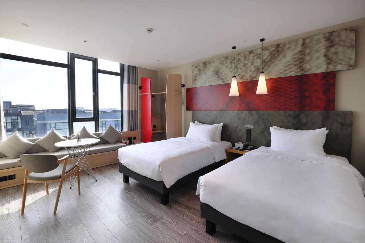 A hotel room with two beds, a seating area with a table, and a large window offering a city view.