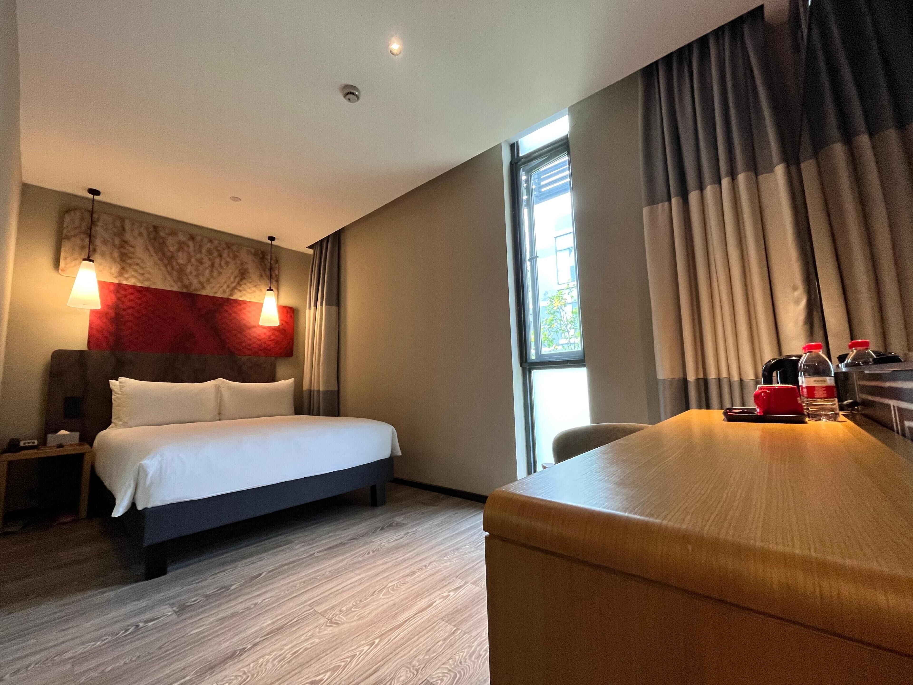 A hotel room with a bed, bedside table, lamp, curtains, and a desk with a chair.