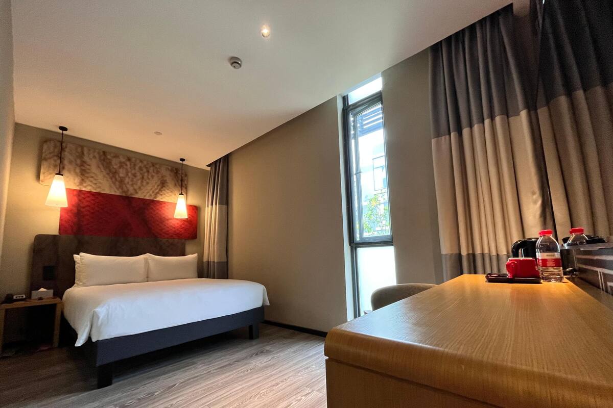 A hotel room with a bed, bedside table, lamp, curtains, and a desk with a chair.