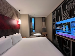 A modern hotel room with a large bed, a TV on the wall, a small table with a lamp, and a view of the city through the window.