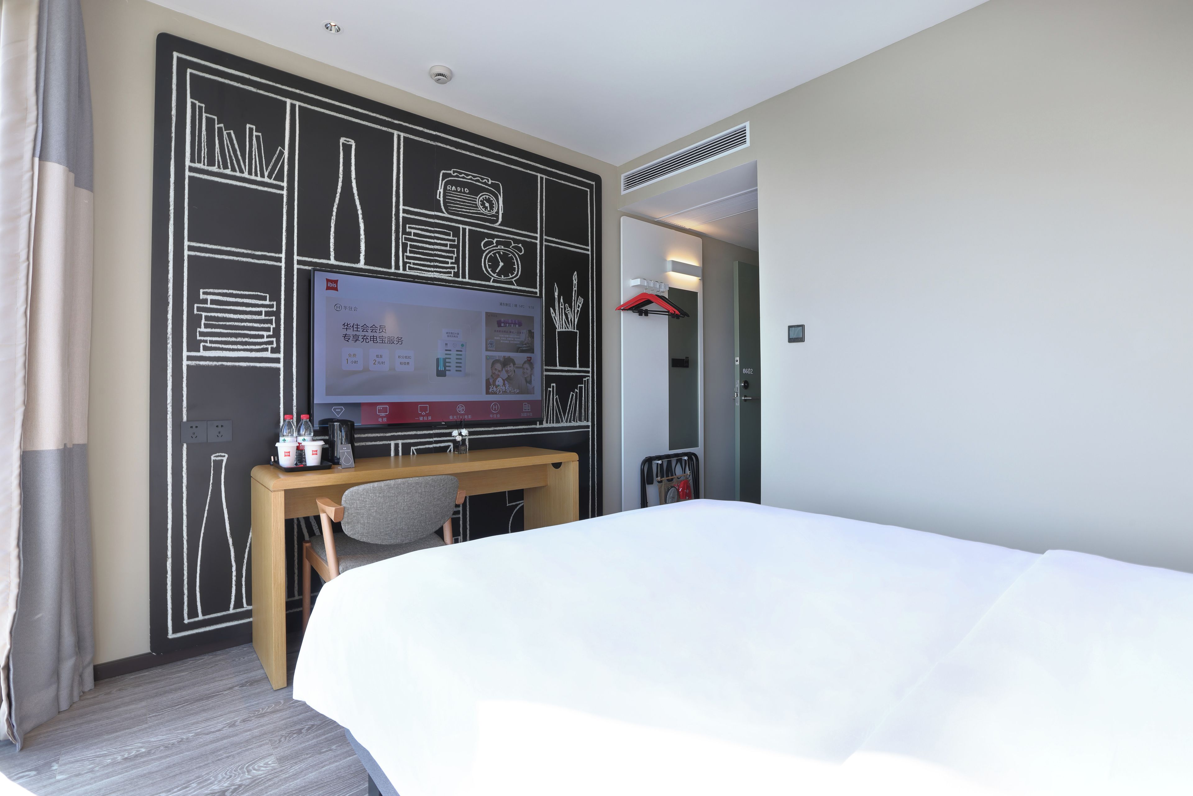 A hotel room with a bed, a desk with a computer, a television, and a wall with a chalkboard design.