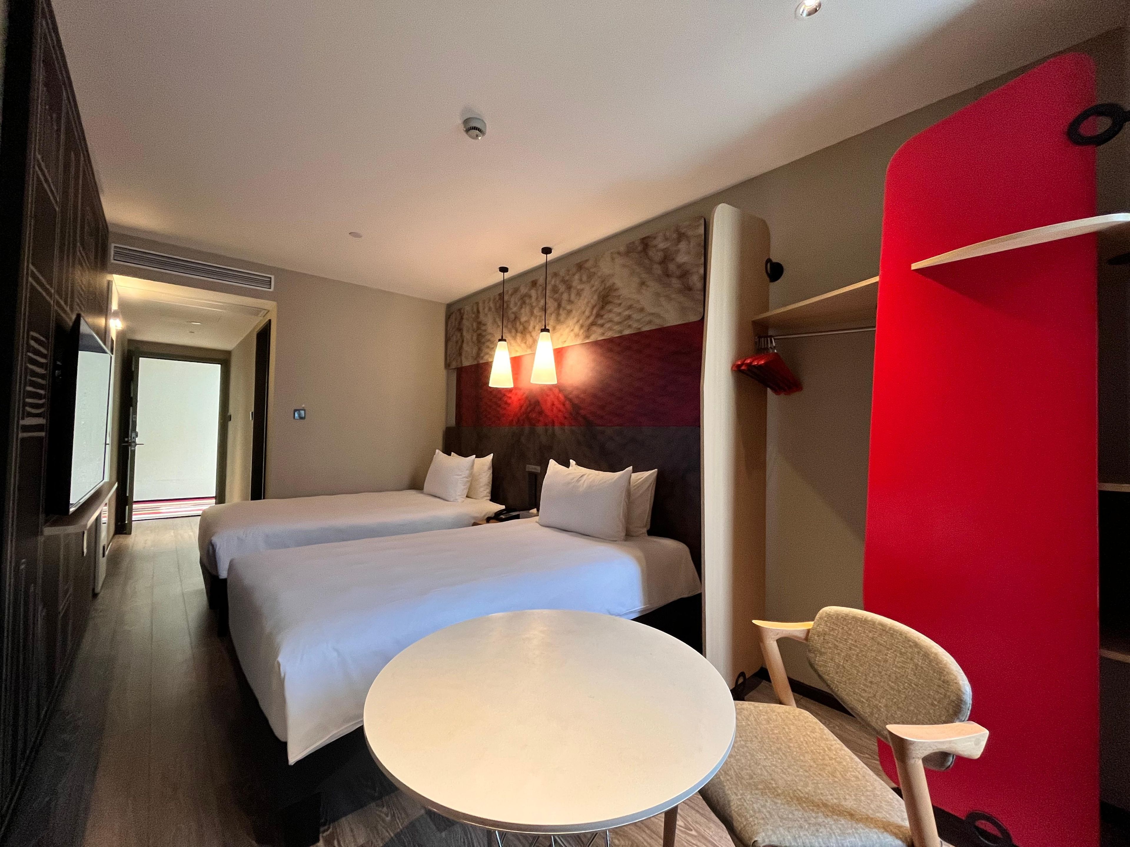 A hotel room with two beds, a round table, a chair, and a red accent wall.