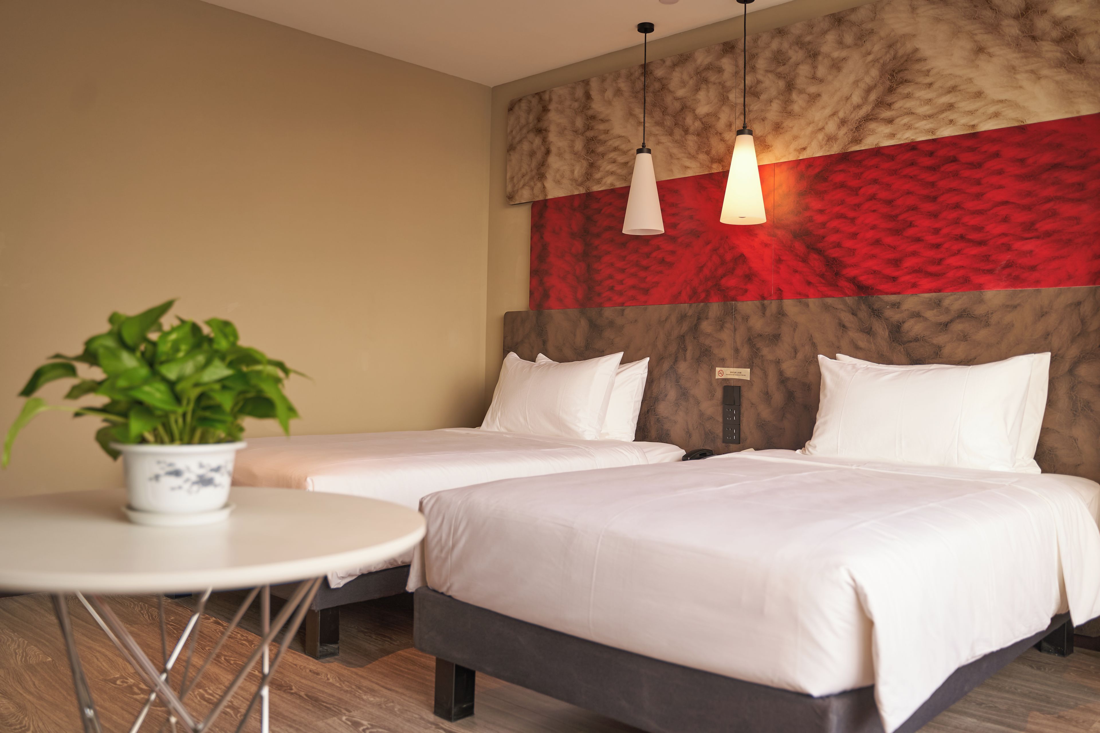 A hotel room with two beds, a small round table with a potted plant, and a red and beige textured wall.