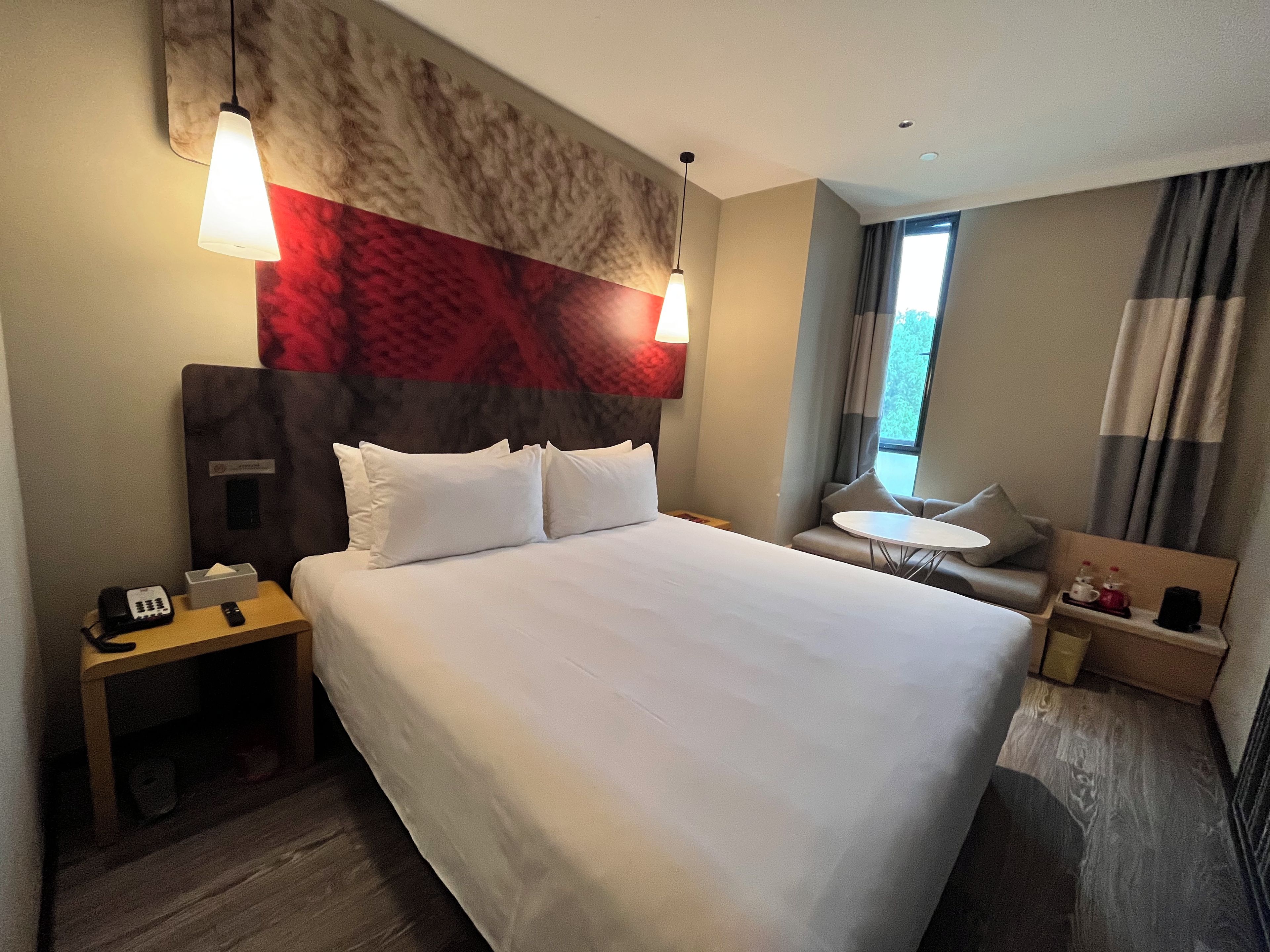 A hotel room with a large bed, bedside tables, a small round table, a sofa, and a window with curtains.